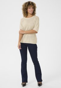 Haut en tricot beige texturé avec des manches larges, associé à un jean évasé bleu foncé. Le modèle porte des escarpins noirs brillants à bout pointu.