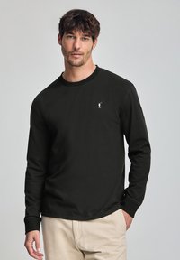 Svart långärmad t-shirt i bomull, med en ribbad rundhals och en liten vit logotyp på vänster bröst. Slät textur.