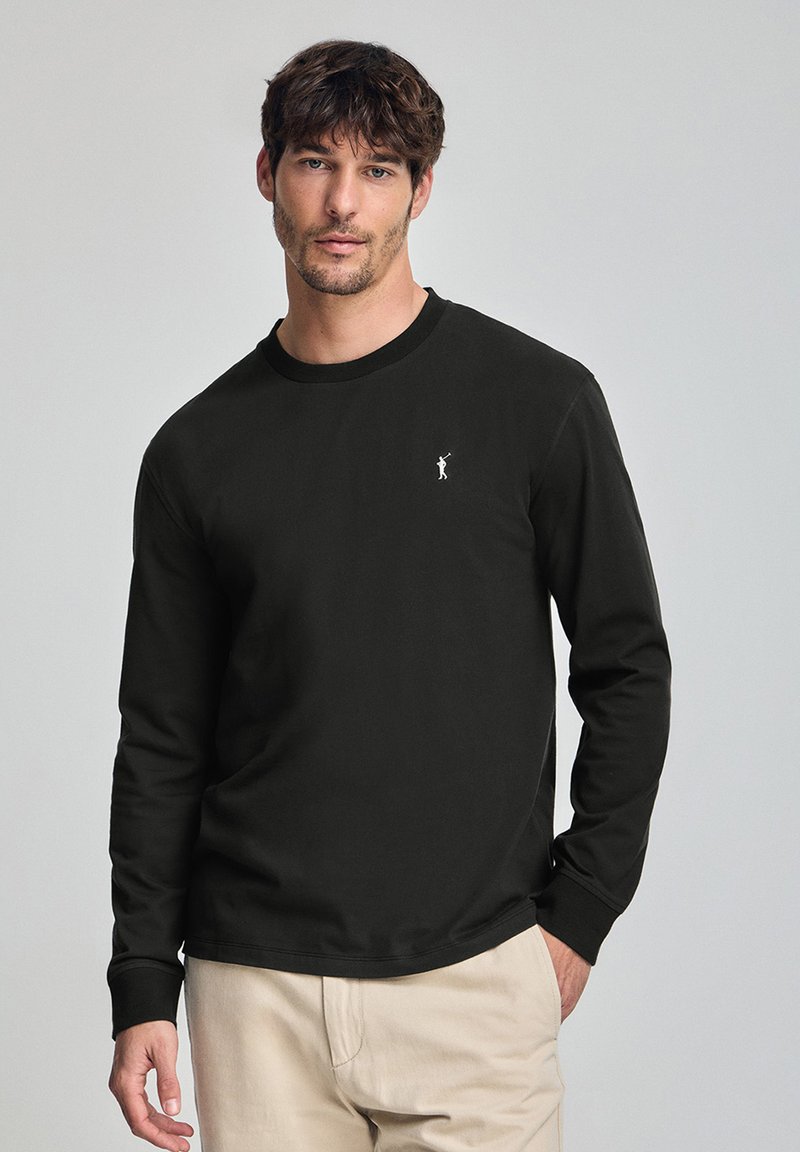 Svart långärmad t-shirt i bomull, med en ribbad rundhals och en liten vit logotyp på vänster bröst. Slät textur.