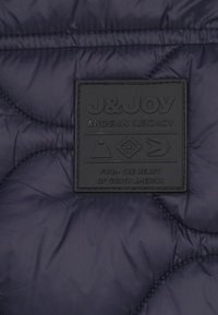 Marinegequilte pufferstof met een gestructureerd zwart rubberen label. Het label bevat de tekst "J&JOY ANDEAN LEGACY" en geometrische symbolen.