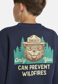 Marineblauwe sweater met een afbeelding van een beer die een rangerhoed draagt, omringd door groene bomen. De tekst luidt: "Jij alleen kunt bosbranden voorkomen."