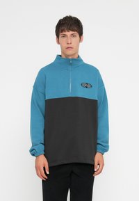 Fargeblokkert fleece-pull i teal og svart med høy krage, halv-glidelås og brodert logo på brystet. Elastiske mansjetter.