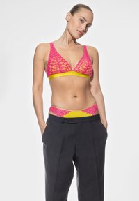 Sostén triangular inalámbrico en encaje neón rosa con banda amarilla; cinturilla de encaje a juego asomando bajo pantalones sueltos grises carbón.