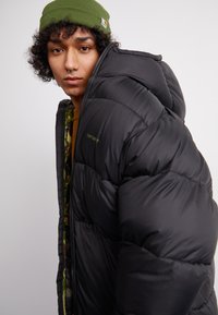 Schwarze Puffjacke mit gestepptem Design, sichtbaren Nähten und einem grünen Innenfutter. Verfügt über einen Reißverschluss und eine Kapuze. Mit einem Logo auf der Brust akzentuiert.