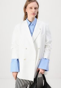 White double-breasted blazer, blue button-up shirt with larger cuffs, black striped pants, and a black handbag. Smooth, tailored fabric.