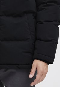 Black puffer jacket with a smooth surface, quilted sections, elastic cuffs, and a side pocket. Hand resting on the fabric.