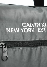 Bolso de nylon gris con un bolsillo con cremallera, "CALVIN KLEIN NEW YORK EST" en texto blanco y un pequeño logotipo negro en el tirador de la cremallera.