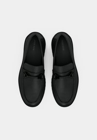 Μαύρα δερμάτινα μοκασίνια με λεία υφή, που διαθέτουν μια διακριτική υφασμένη λεπτομέρεια και μαύρο μεταλλικό διακοσμητικό στοιχείο στο πάνω μέρος.