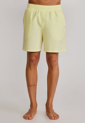 Man in lichtgele shorts met elastische taille, staand op blote voeten tegen een eenvoudige neutrale achtergrond.