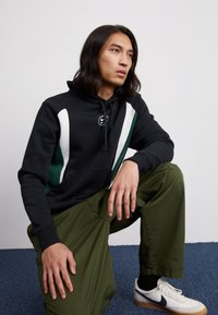 Mustat huppari valkoisilla ja vihreillä yksityiskohdilla, jossa on nyöritettävä huppu ja logo. Yhdistetty oliivinvihreisiin housuihin ja valkoisiin tennareihin.