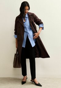 Cappotto trench marrone sopra una camicia azzurra chiara a bottoni, pantaloni neri e décolleté neri a punta. È visibile anche una borsa marrone.