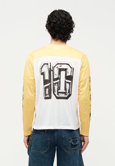 Άντρας με σγουρά μαλλιά που φοράει κίτρινο-άσπρο μακρυμάνικο πουκάμισο με μεγάλο φθαρμένο αριθμό "10" στην πλάτη και μπλε τζιν, φαίνεται από πίσω.