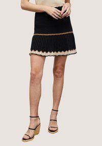 Stickad minikjol i svart med vågiga kanter i beige och brunt. Kombinerad med svarta sandaler med remmar och kilklackar på en ljus bakgrund.