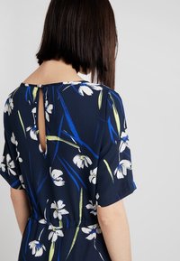 Kukkakirjailtu mekko tummansinisestä kankaasta, jossa on takana avainaukko, lyhyet hihat ja kapeampi vyötärö, jossa on valkoisia ja vihreitä kukkakuoseja.