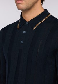 Polo shirt blu navy con texture a coste verticali, con colletto a righe marroni in contrasto e bottoni blu.