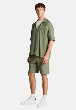 Młody mężczyzna stoi, ubrany w jasnooliwkową koszulę na krótki rękaw na guziki, pasujące szorty, białe skarpetki i białe trampki na jasnym tle.