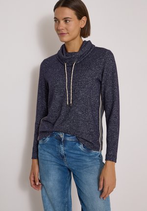Femme portant un pull à col boule bleu foncé avec des cordons et un jean bleu clair, debout devant un fond clair uni.