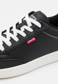 Baskets synthétiques noires avec des lacets blancs, arborant un logo Levi's rouge, une texture perforée sur les côtés, et une semelle en caoutchouc blanche.