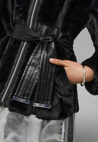 Schwarze Fake-Fur-Jacke mit tailliertem Schnitt, Lederakzenten, Reißverschluss und silbernen Details, mit einer Hand in der Tasche zur Größeneinschätzung.