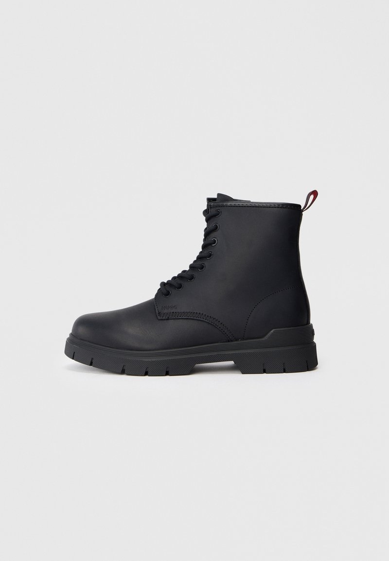 Bottine noire en cuir avec une semelle épaisse, devant à lacets et une languette rouge sur un fond clair uni.