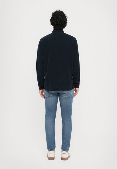 Cămașă de un albastru închis, cu mâneci lungi, guler și manșete cu nasturi, purtată cu blugi slim-fit de culoare albastru deschis și încălțăminte casual.
