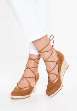 Benen in witte broek met tan suede wedge espadrilles met gekruiste enkelbandjes die boven de enkels zijn geknoopt op een witte achtergrond.