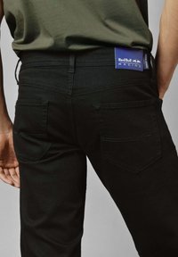 Takaisin näkymä henkilöstä, joka on pukeutunut mustiin farkkuihin, joissa on Red Bull Racing -patchi, ja oliivinvihreään paitoihin, käsi rentona sivulla.