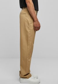 Beige chinos with a straight cut design, featuring a back pocket and textured fabric. Worn with white sneakers for a casual look.