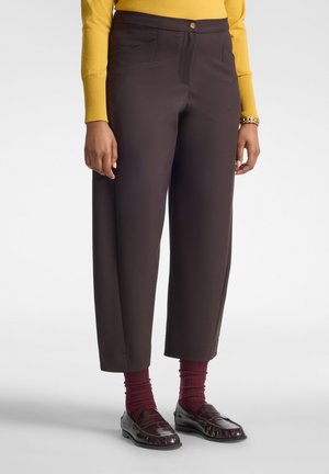 Person, die dunkelbraune, verkürzte Hosen, kastanienbraune Socken, dunkelbraune Loafers und ein gelbes Langarmshirt trägt, vor einem einfarbigen Hintergrund steht.