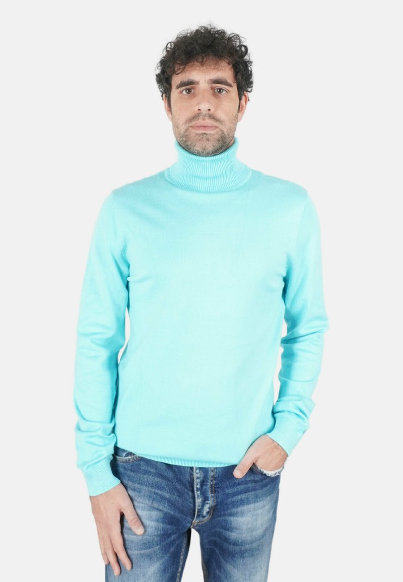 Uomo con capelli ricci che indossa un dolcevita azzurro chiaro e jeans blu, in piedi con una mano in tasca su sfondo bianco.