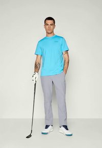 Kék inget és szürke nadrágot viselő férfi, aki egy golfütőt tart, bal kezén fehér kesztyűvel, egy világos háttér előtt áll.