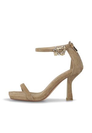 Sandale à talon haute beige avec une finition en daim. Présente une fine bande autour de la cheville, une large bande au niveau des orteils et des accents décoratifs en strass.