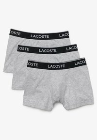 Boxer-briefs en coton gris, avec une ceinture élastique noire portant le logo "LACOSTE" en blanc. Trois paires empilées, texture lisse.