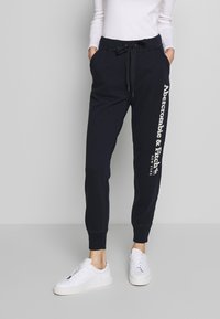 Vrouw in zwarte Abercrombie & Fitch sweatpants met trekkoord en witte sneakers, staande tegen een effen lichtgrijze achtergrond.