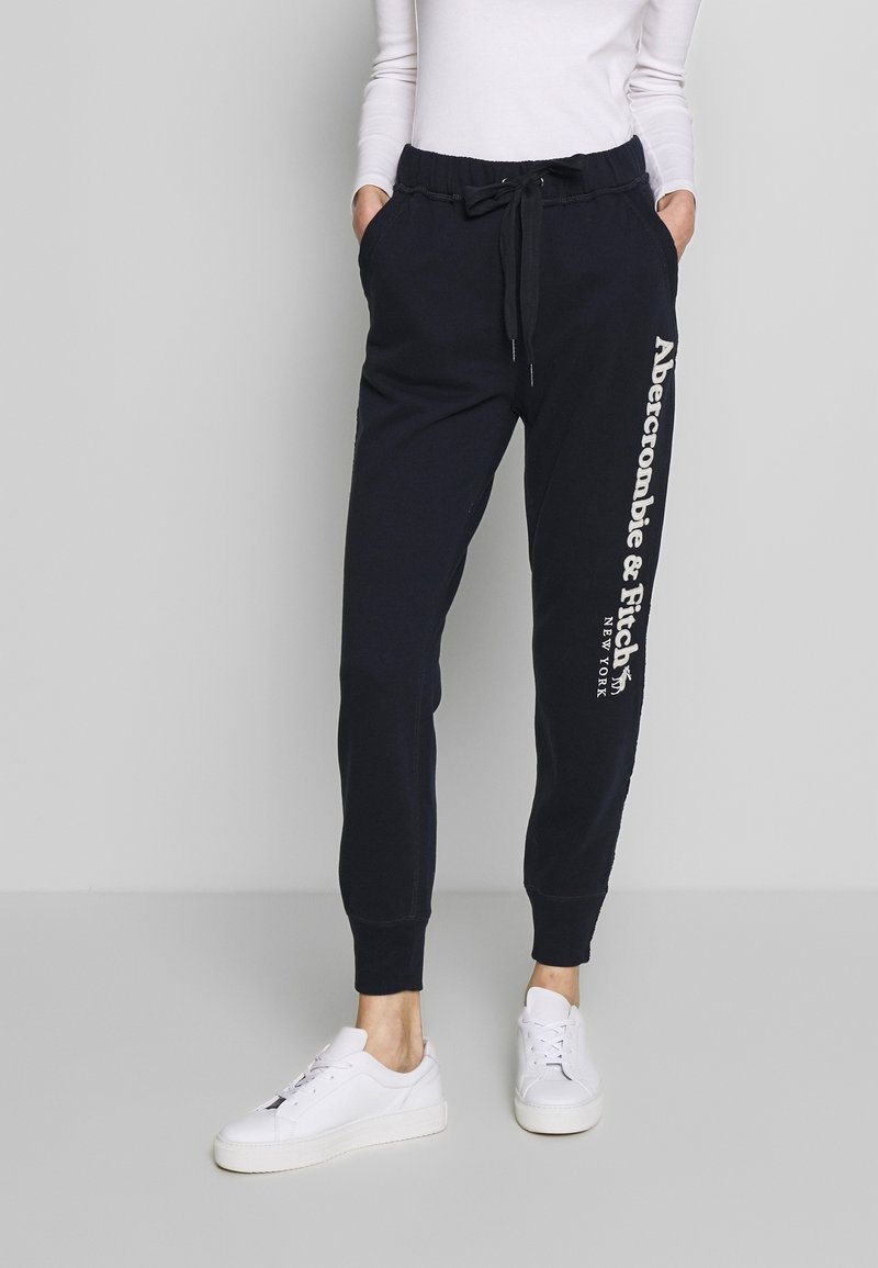 Vrouw in zwarte Abercrombie & Fitch sweatpants met trekkoord en witte sneakers, staande tegen een effen lichtgrijze achtergrond.
