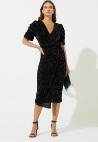 Svart paljettklänning med djup V-ringning, puffärmar och figursydd midja. Knälång, mjuk textur, ihop med svarta ankelremsskor och en kuvertväska.