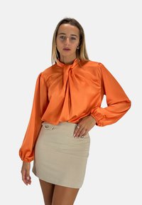 Oranje satijnen blouse met lange ballonmouwen en een strikje bij de halslijn, gecombineerd met een beige mini-jurk met twee knopen aan de voorkant.