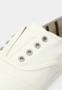 Chaussure en toile blanche avec des œillets argentés et un motif cousu. La zone des orteils est arrondie et l'intérieur présente un motif rayé.