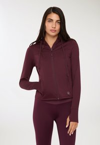 Donna che indossa una felpa aderente bordeaux con zip e fori per il pollice, abbinata a leggings, in piedi con una mano in tasca su uno sfondo bianco.