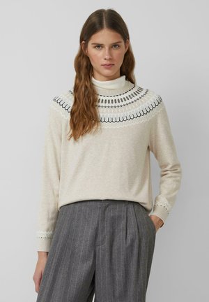 Un pull beige à col montant, orné de broderies noires délicates autour du col. Manches longues avec des poignets décoratifs. Pantalons à rayures grises.