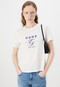T-shirt in cotone bianco con stampa grafica blu navy "GANT EST. 1949 NEW HAVEN." Maniche corte, vestibilità rilassata, abbinata a jeans in denim azzurro chiaro e una borsa nera.