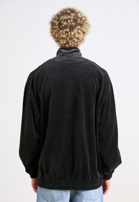 Svart, fløyelsjakke med høy krave, elastisk kant og senket skuldre. Har en myk tekstur og avslappet passform.
