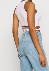Top corto blanco con estampado de tie-dye en rosa y amarillo; presenta un escote abierto con tirantes entrecruzados. Combinado con unos vaqueros de tiro alto en azul claro.