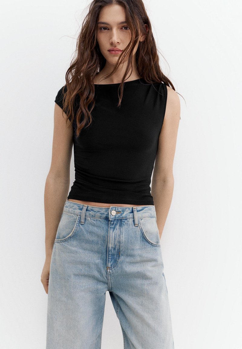 Junge Frau mit langen braunen Haaren, die ein eng anliegendes schwarzes ärmelloses Oberteil und hellblaue hoch taillierte Jeans trägt, steht vor einem weißen Hintergrund.