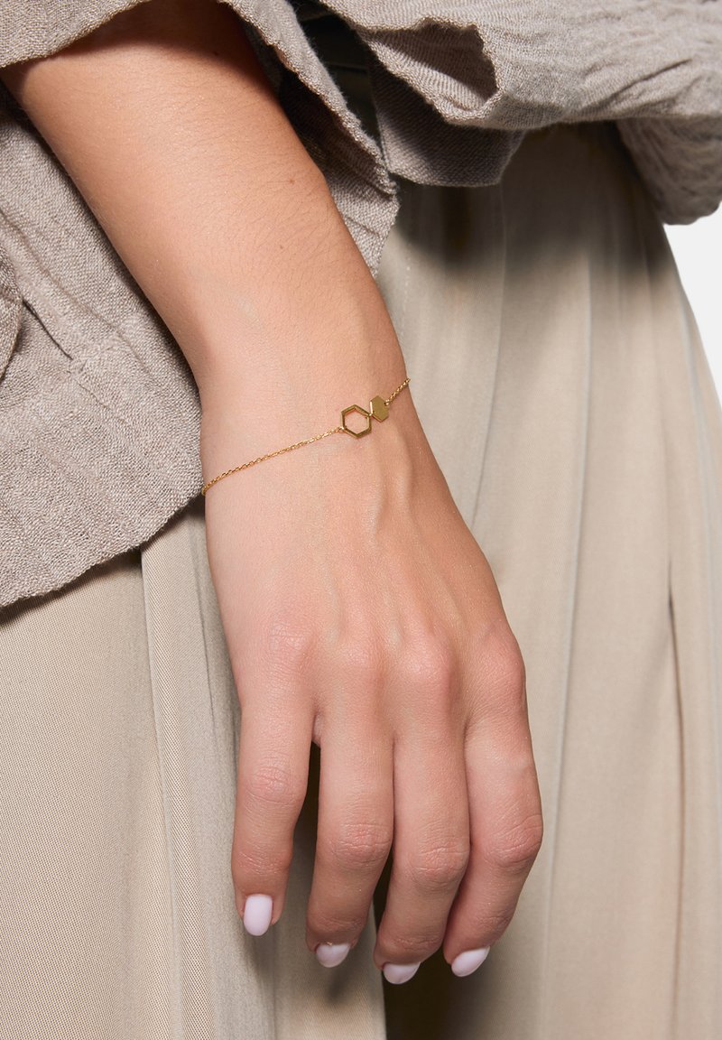 Złota bransoletka łańcuszkowa z detalem w kształcie geometrycznego sześciokąta i serce, noszona na dłoni na tle miękkiego, fakturowanego materiału.