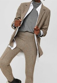 Geruit colbert in beige en bruin met bijpassende, nauwsluitende broek. Grijze coltrui eronder, daarboven een wit overhemd zichtbaar. Zwarte laarzen.