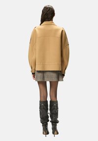 Veste en laine beige surdimensionnée à épaules tombantes, associée à une jupe à carreaux et des bottes en cuir noir montantes jusqu'aux genoux, vue de dos.