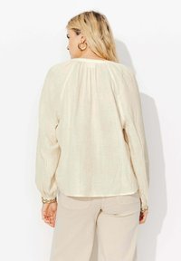 Blouse beige clair à manches longues avec un design fluide, texture de tissu douce, dos froncé et quincaillerie dorée sur les poignets.