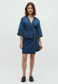 Jesionka z denimu z szerokimi rękawami i wiązaniem, zestawiona z dopasowaną mini spódniczką z denimu. Oba elementy w ciemnoniebieskim odcieniu. Gładka tekstura.