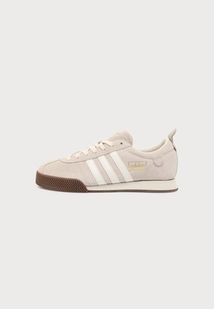 Béžová teniska Adidas s bielymi pásikmi, zlatým logom, béžovým semišovým zvrškom, bielymi šnúrkami a hnedou gumovou podrážkou na jednoduchom pozadí.